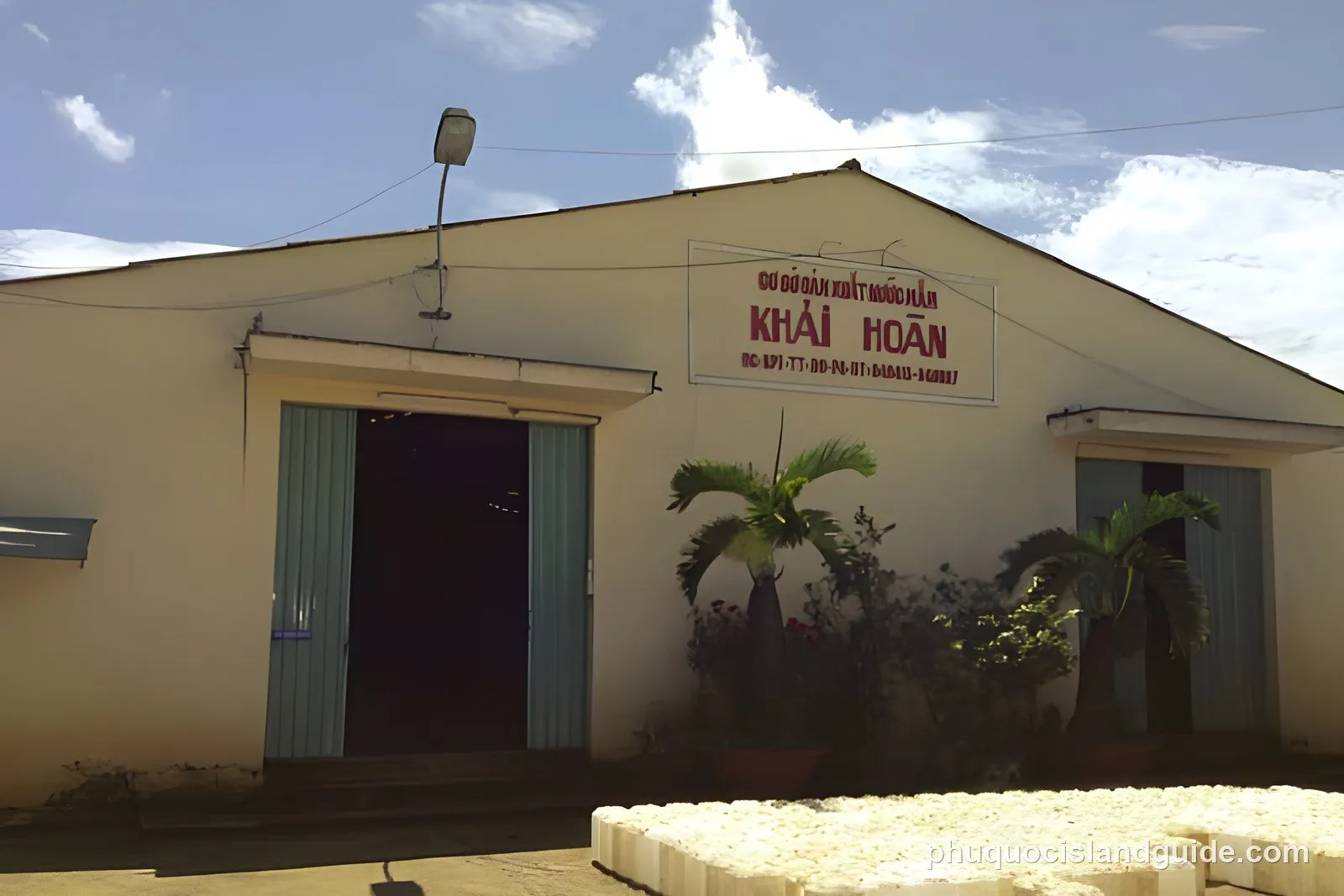 Exterior view of a building with a sign in Vietnamese that reads ‘CƠ SỞ SẢN XUẤT NƯỚC MẮM KHẢI HOÀN’, tropical plants, and stacked white blocks.