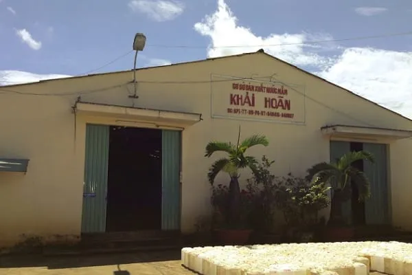 Exterior view of a building with a sign in Vietnamese that reads ‘CƠ SỞ SẢN XUẤT NƯỚC MẮM KHẢI HOÀN’, tropical plants, and stacked white blocks.
