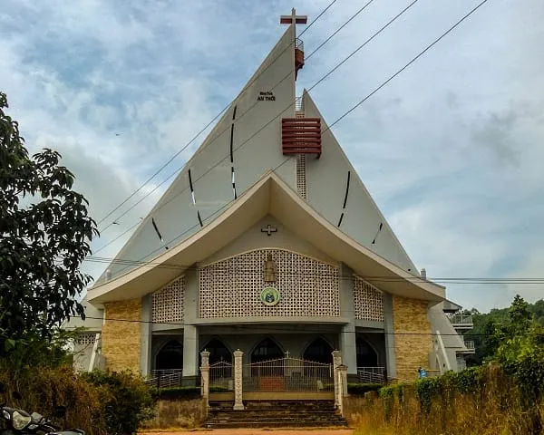 Front view of a modern church with a triangular roof, a cross on top, and a patterned facade, surrounded by green tropical trees under a cloudy sky.