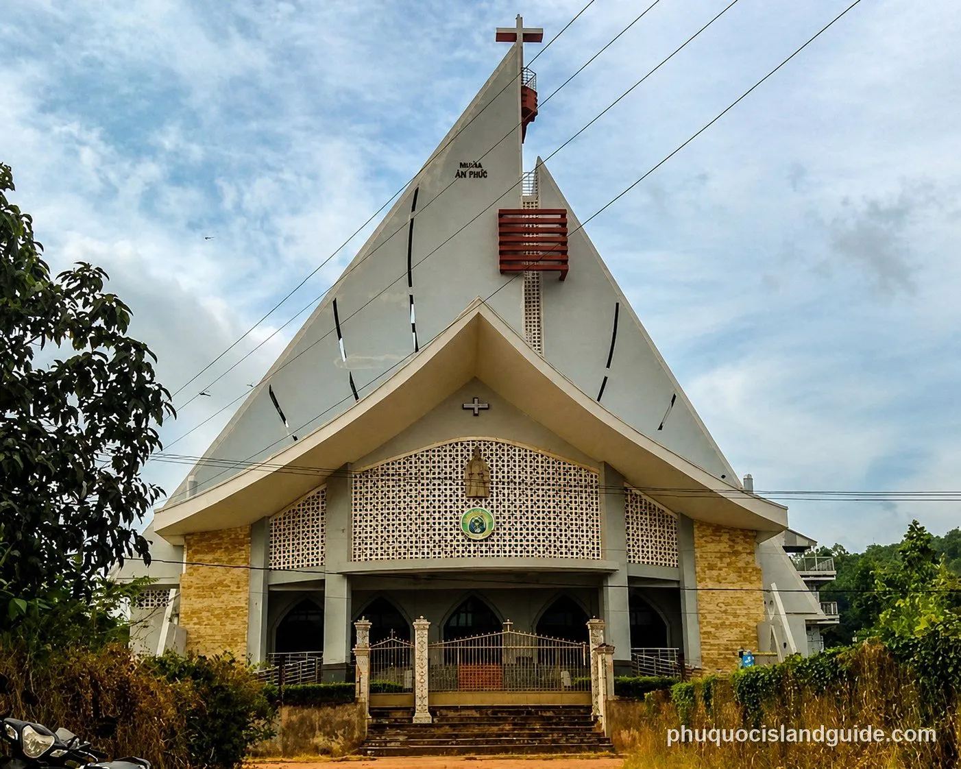 Front view of a modern church with a triangular roof, a cross on top, and a patterned facade, surrounded by green tropical trees under a cloudy sky.