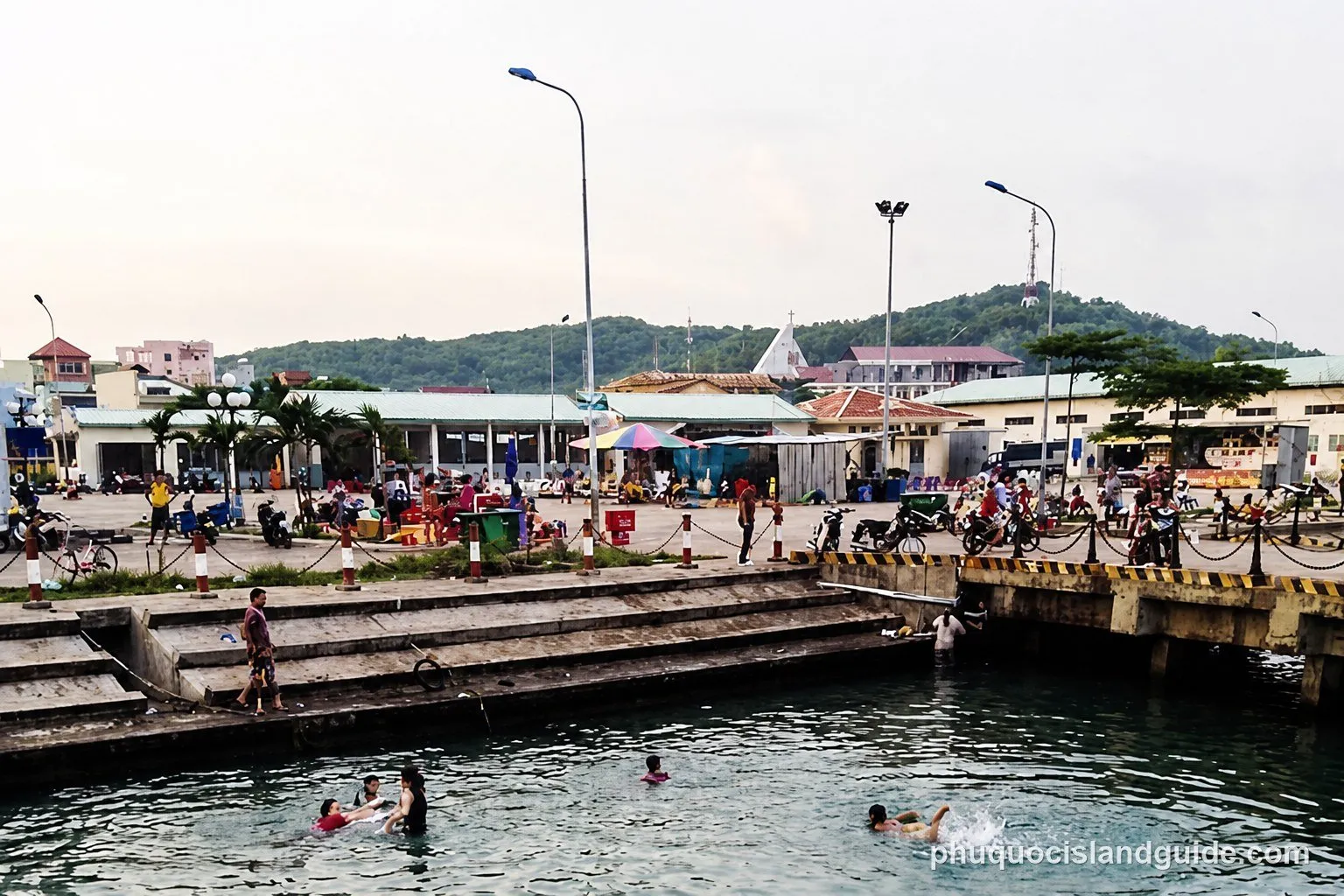 A bustling waterfront scene with people swimming in the foreground, buildings, and a green, forested hill in the background.