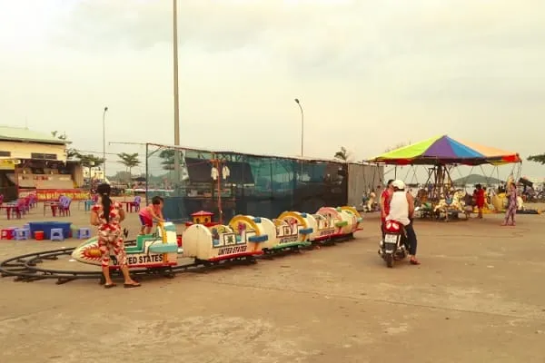 An outdoor amusement area with a kiddie train ride and a colorful carousel under a cloudy sky. People are present, with buildings in the background.