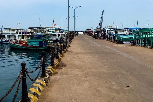 A busy concrete pier with several fishing and passenger boats docked on both sides. People are visible on the pier, and a crane stands in the background.