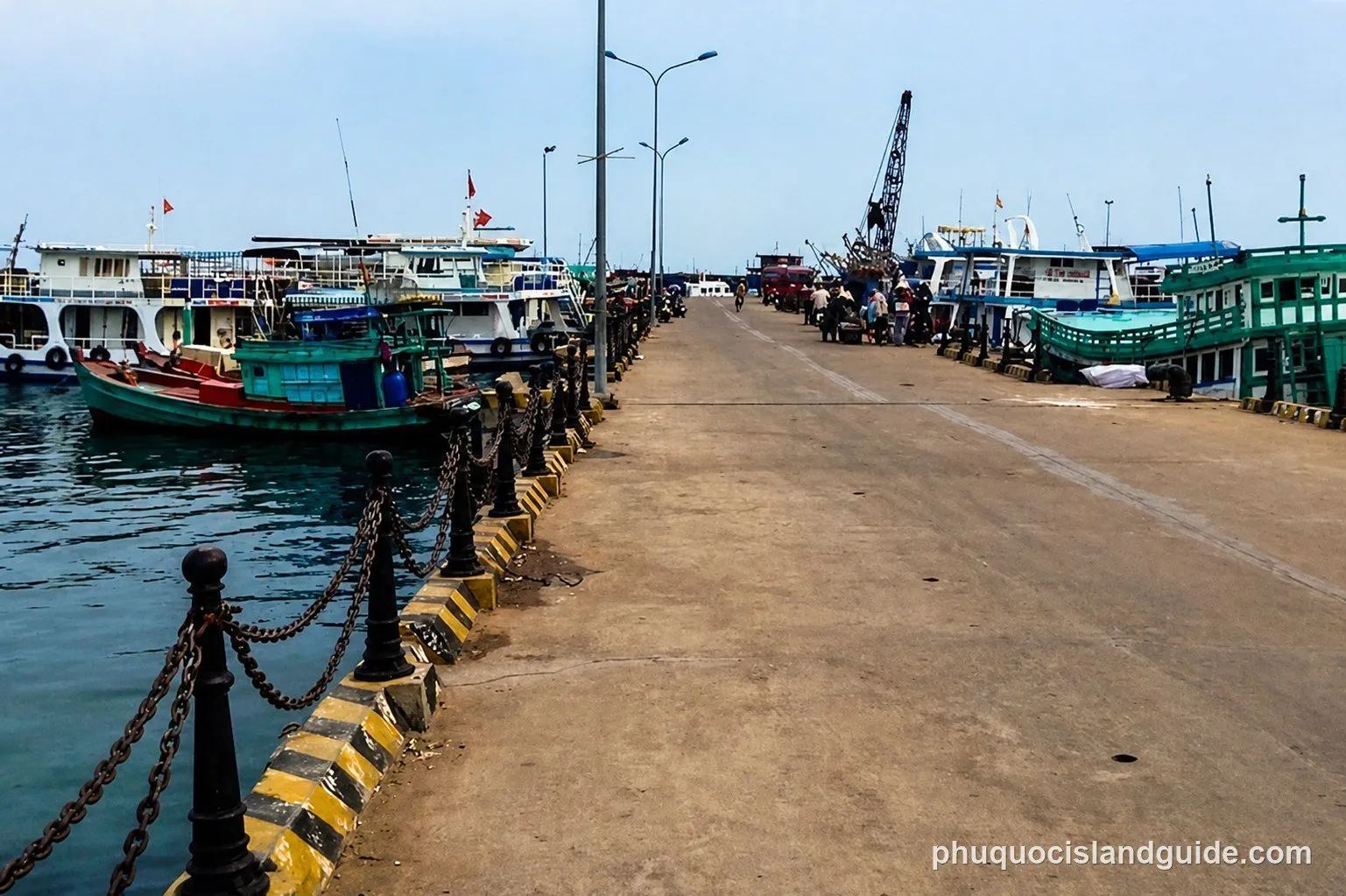 A busy concrete pier with several fishing and passenger boats docked on both sides. People are visible on the pier, and a crane stands in the background.