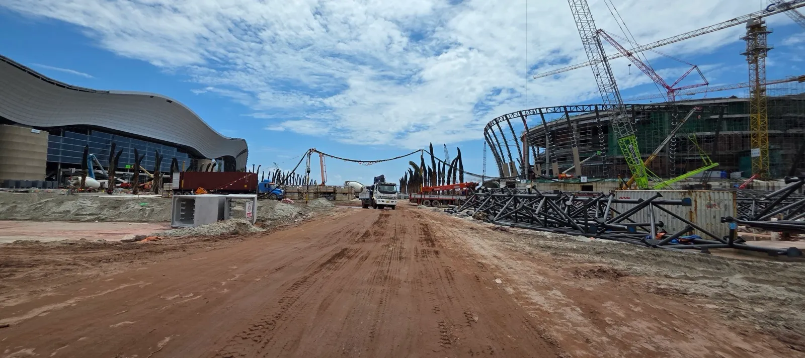 The finished wave-shaped roof of the convention centre on the left and the steel frame of the round performance hall with cranes on the right, either side of a dirt road