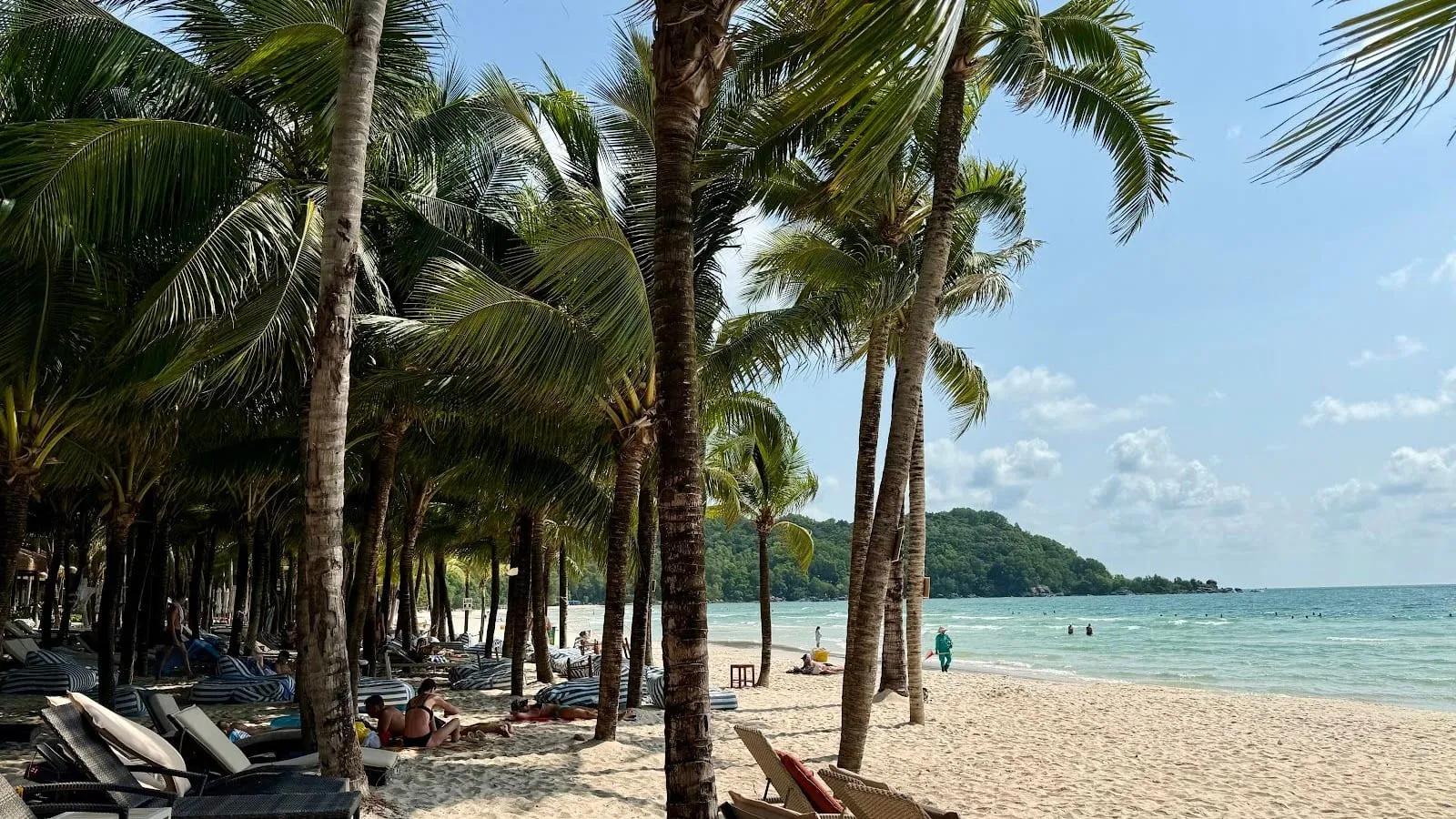 Palm trees and sunbeds on the white sand of Khem Beach, with the sea and a wooded headland behind, in April