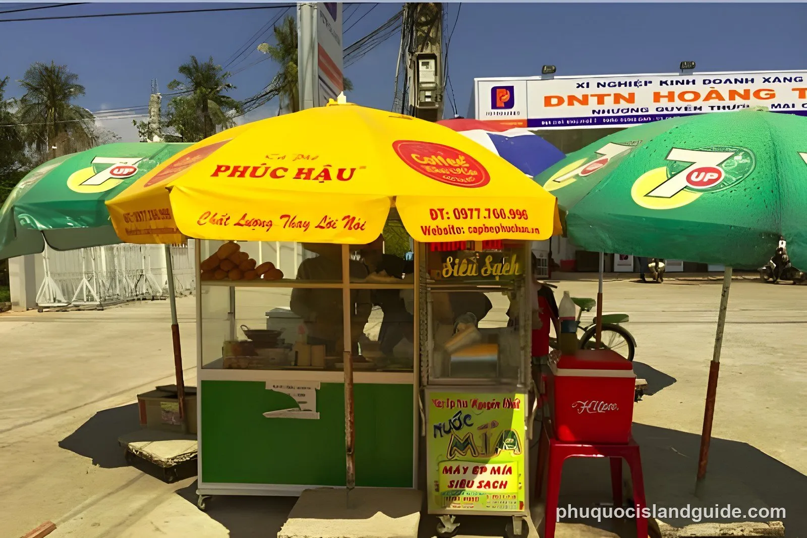 A street vendor stall with yellow and green umbrellas selling coffee and sugarcane juice on a sunny day in a tropical setting. A large billboard is in the background.