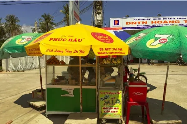 A street vendor stall with yellow and green umbrellas selling coffee and sugarcane juice on a sunny day in a tropical setting. A large billboard is in the background.