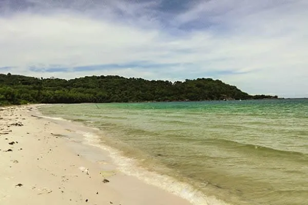 A scenic view of a sandy beach with clear turquoise water and a lush green, forested island in the background under a cloudy sky.