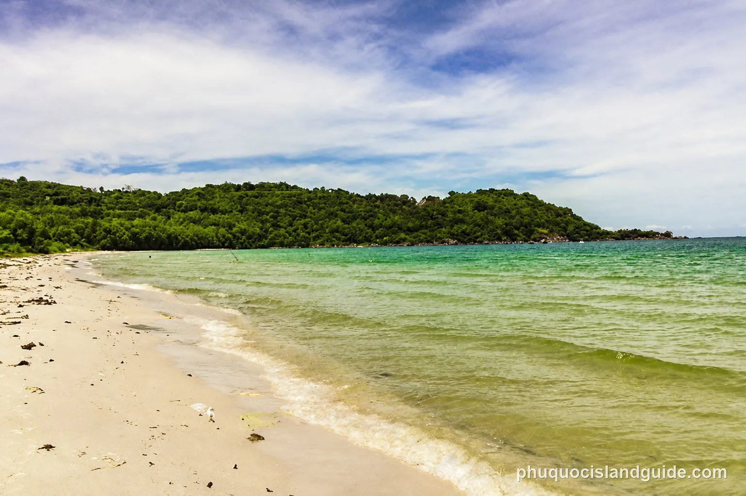 A scenic view of a sandy beach with clear turquoise water and a lush green, forested island in the background under a cloudy sky.