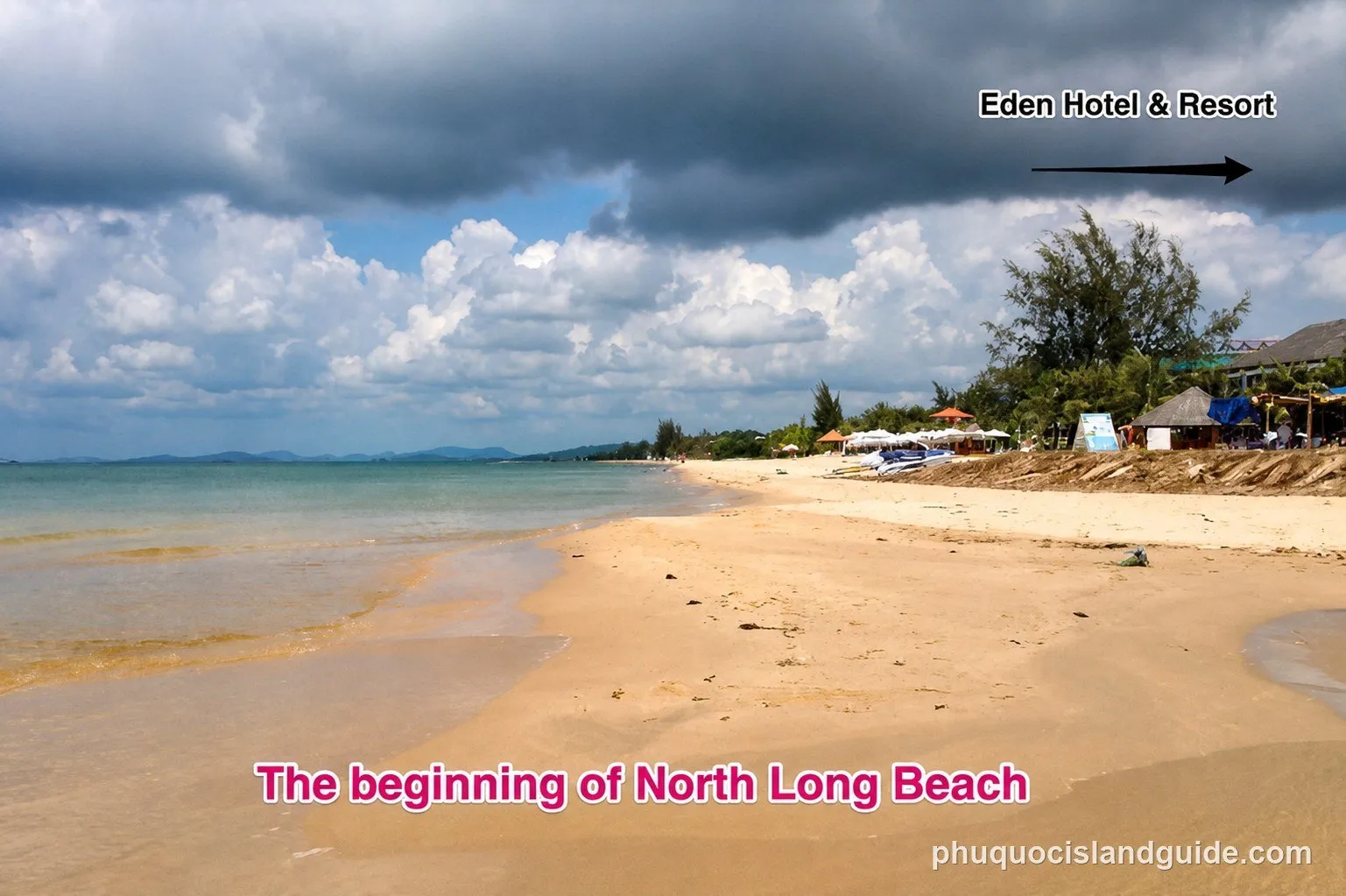 A wide shot of a sandy beach with calm ocean water under a cloudy sky. Buildings and tropical trees line the right side of the beach.