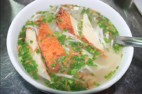 A close-up shot of a bowl of Vietnamese fish noodle soup (bún cá) with clear broth, white noodles, fried fish cakes, and chopped green onions, with a spoon.