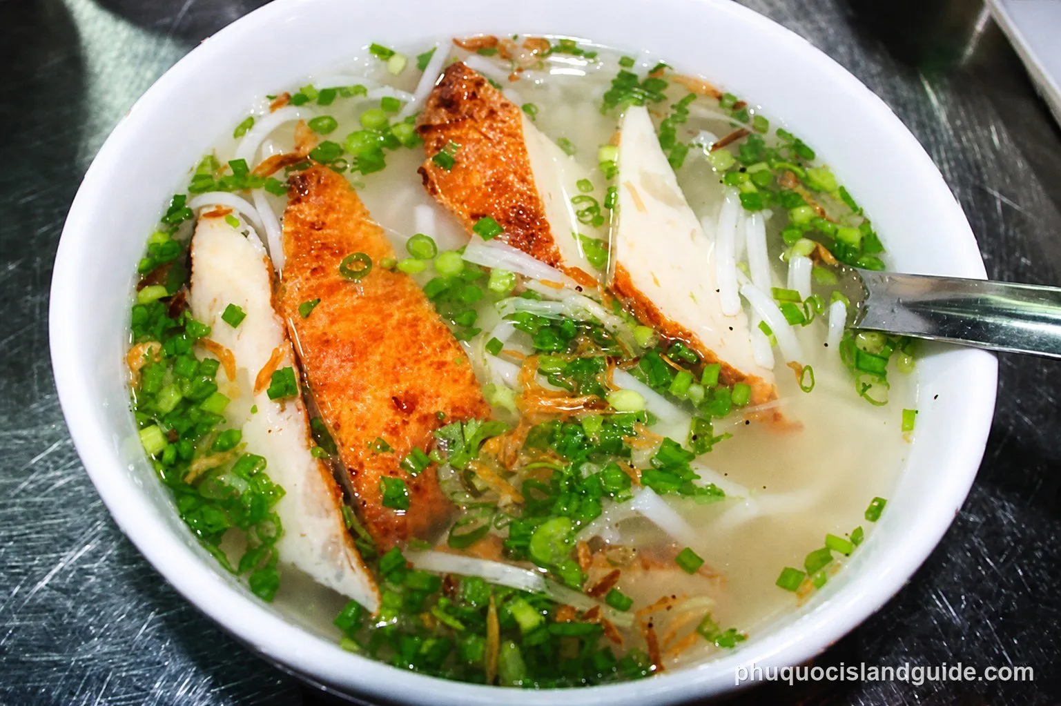 A close-up shot of a bowl of Vietnamese fish noodle soup (bún cá) with clear broth, white noodles, fried fish cakes, and chopped green onions, with a spoon.