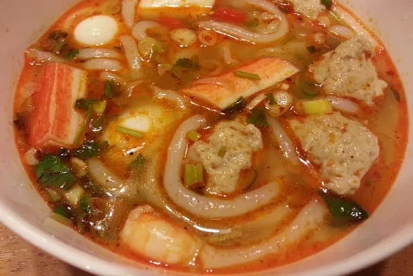 A close-up of a bowl of Vietnamese noodle soup with crab sticks, fish balls, quail eggs, shrimp, and green onions in a spicy, reddish-orange broth.