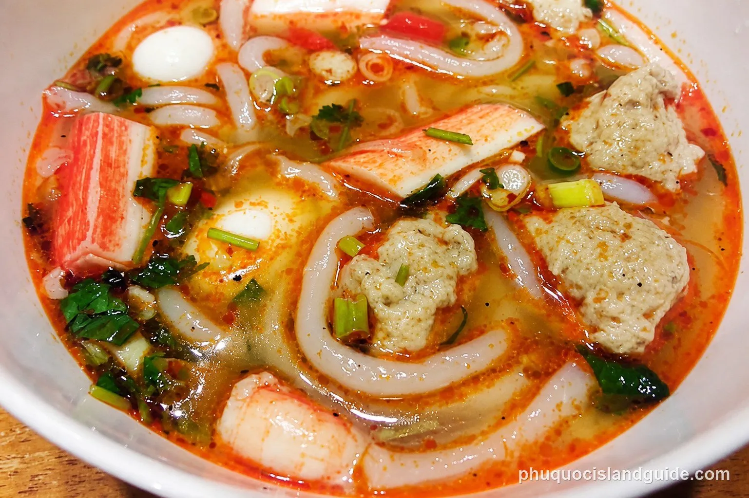 A close-up of a bowl of Vietnamese noodle soup with crab sticks, fish balls, quail eggs, shrimp, and green onions in a spicy, reddish-orange broth.