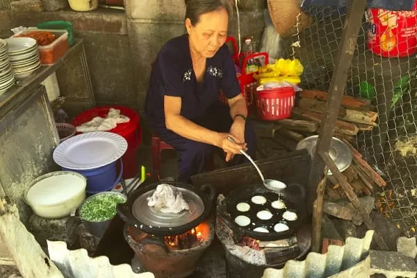 An elderly Vietnamese woman cooking small pancakes (bánh khọt) on a street food stall with two charcoal stoves.