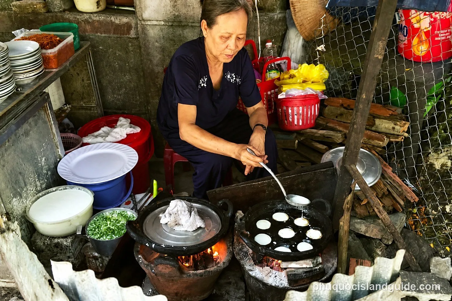 An elderly Vietnamese woman cooking small pancakes (bánh khọt) on a street food stall with two charcoal stoves.