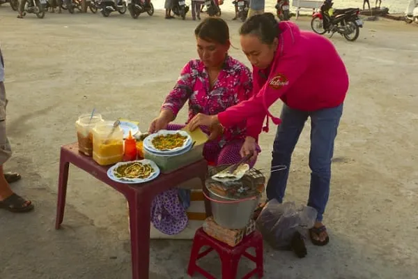 Two women prepare and serve street food from a small table and charcoal grill on a paved area, with motorbikes parked in the background.