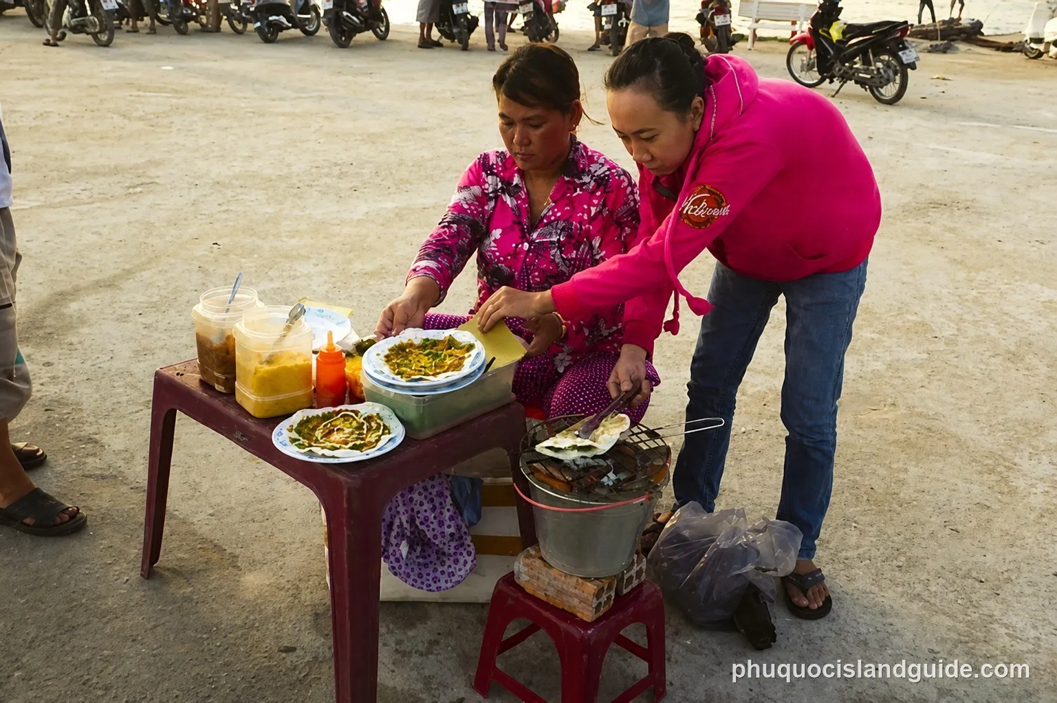 Two women prepare and serve street food from a small table and charcoal grill on a paved area, with motorbikes parked in the background.