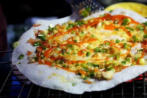 A close-up shot of a Vietnamese grilled rice paper pizza (Bánh tráng nướng) with green onions, minced meat, and chili sauce cooking on a charcoal grill.