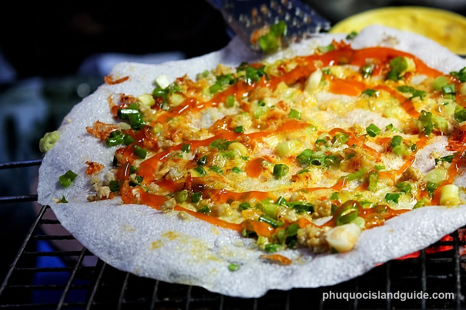 A close-up shot of a Vietnamese grilled rice paper pizza (Bánh tráng nướng) with green onions, minced meat, and chili sauce cooking on a charcoal grill.