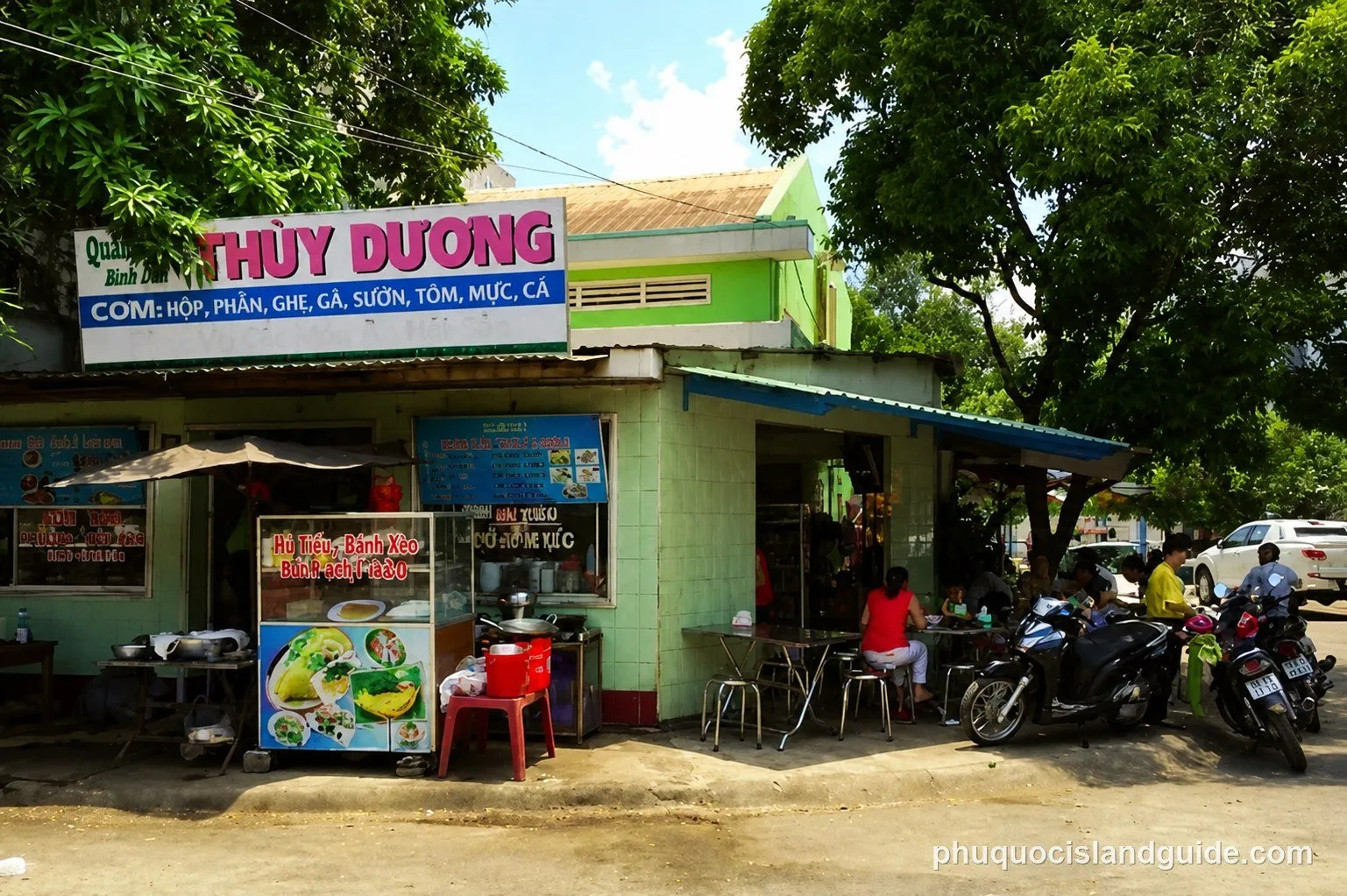 A street-side restaurant in Vietnam with a light green building, a food stall, and people sitting at tables. Motorbikes are parked nearby under green trees.