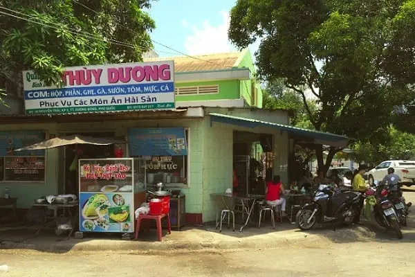 A street-side restaurant in Vietnam with a light green building, a food stall, and people sitting at tables. Motorbikes are parked nearby under green trees.