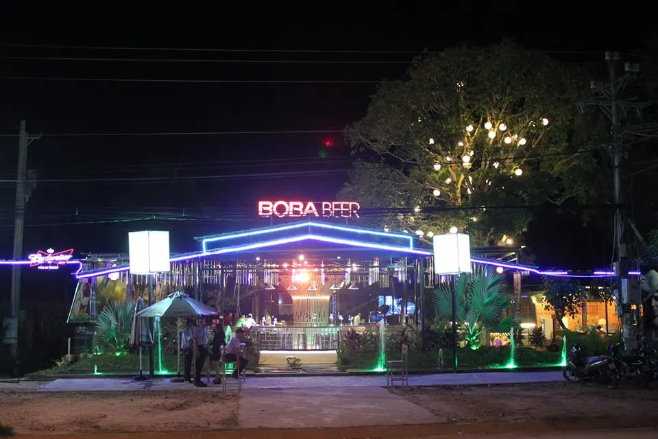 A night photo of a bar or restaurant named “BOBA BEER” with bright neon lights. A large tree decorated with spherical lights stands next to it.