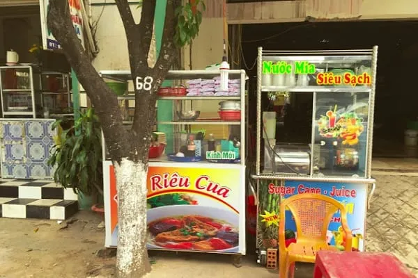 Two street stalls in Vietnam, one selling ‘Rieu Cua’ (crab noodle soup) and the other ‘Nuoc Mia’ (sugarcane juice), with a tree displaying ‘68’ in front.