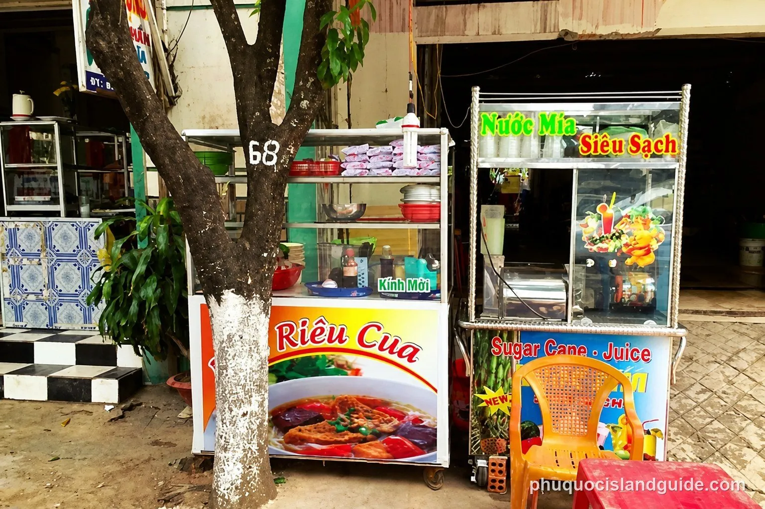 Two street stalls in Vietnam, one selling ‘Rieu Cua’ (crab noodle soup) and the other ‘Nuoc Mia’ (sugarcane juice), with a tree displaying ‘68’ in front.