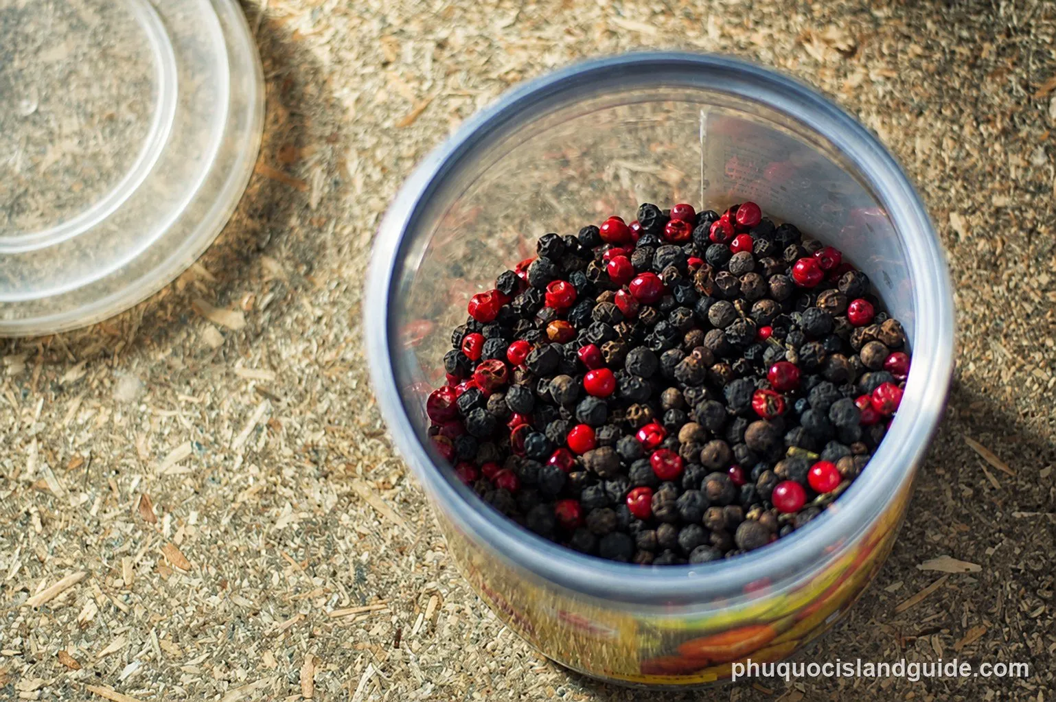 A clear plastic container filled with a mix of black and red peppercorns, with a plastic lid next to it on a textured surface.