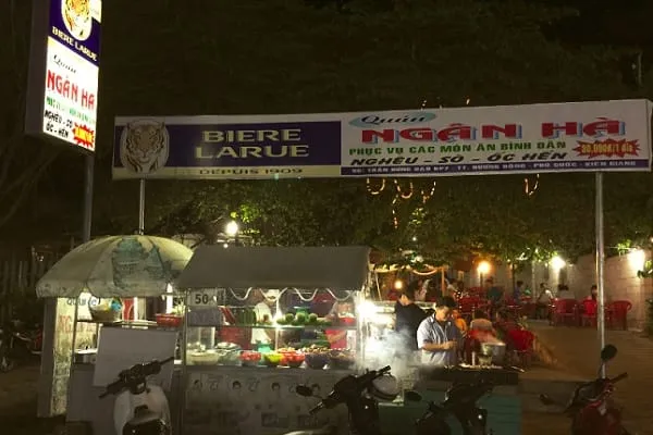 An outdoor food stall and restaurant at night in Vietnam, with illuminated signs for ‘BIERE LARUE’ and ‘QUÁN NGÂN HÀ’, people eating and cooking, and motorbikes.