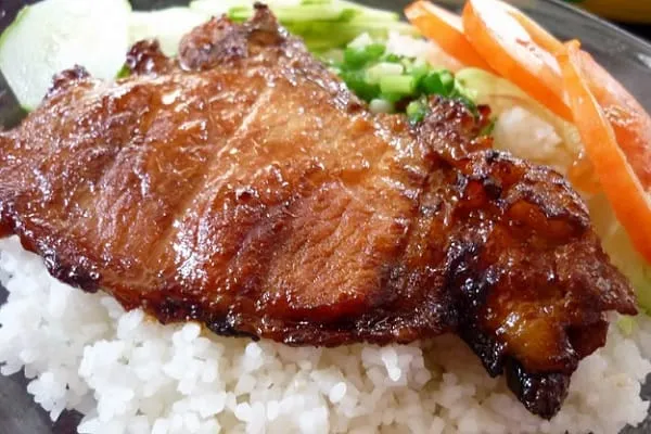 A close-up shot of a plate of Vietnamese broken rice (cơm tấm) with a glistening grilled pork chop, white rice, sliced cucumbers, and tomatoes.