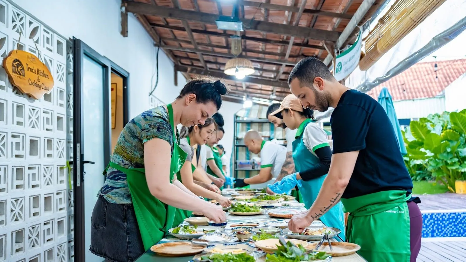 Students in green aprons rolling spring rolls along a long table at Troc's Kitchen cooking class