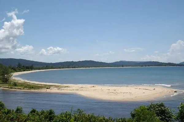 An aerial view of a tropical bay with a wide sandy beach curving along the coastline, meeting a river inlet. Lush green hills and trees surround the bay under a blue sky with white clouds.