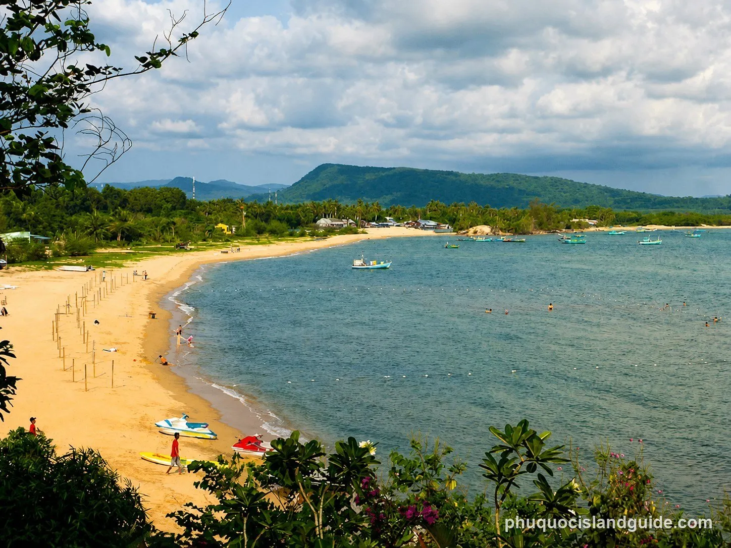 An aerial view of a tropical sandy beach with people swimming and sunbathing, several boats anchored in the clear blue water, and lush green hills in the background.