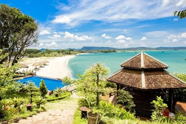 A tropical resort with a swimming pool, a white sandy beach, turquoise ocean, and a traditional tiled gazebo under a clear blue sky.