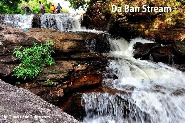 A multi-tiered waterfall cascades over dark rocks, surrounded by green foliage. Several people are visible standing at the top of the waterfall.