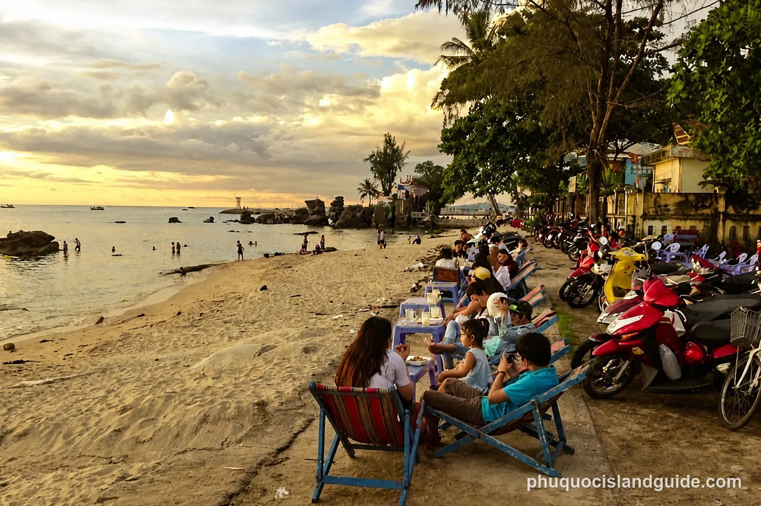 A vibrant sunset over a tropical beach with people relaxing on lounge chairs, swimming in the sea, and motorbikes parked along the sandy shore.
