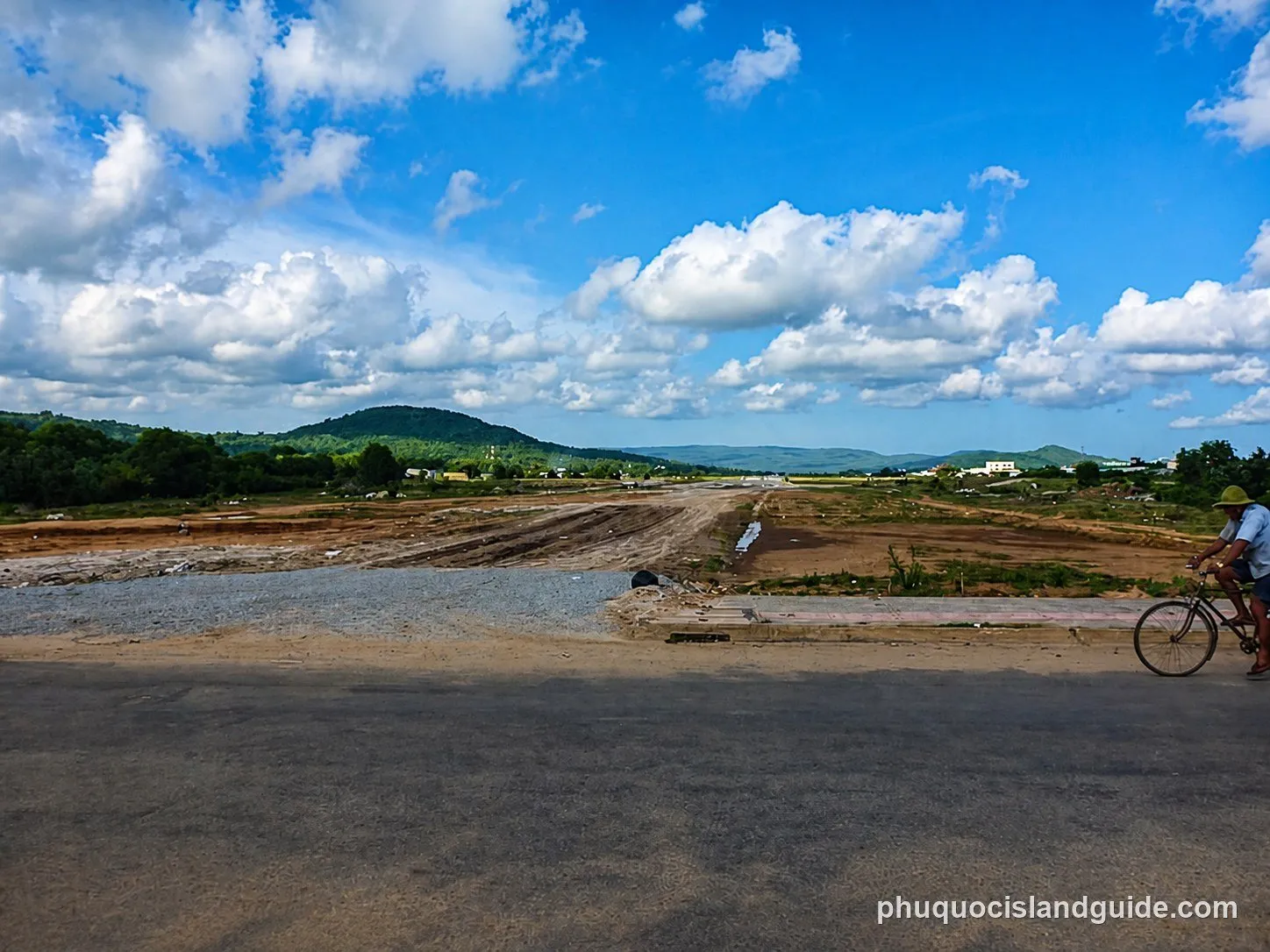 Paved road past cleared land, with green hills behind, in Cua Duong