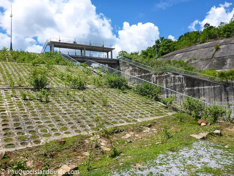 A concrete slope with grass growing through grid patterns, leading up to a concrete structure with stairs and railings. A green hill and blue sky are in the background.