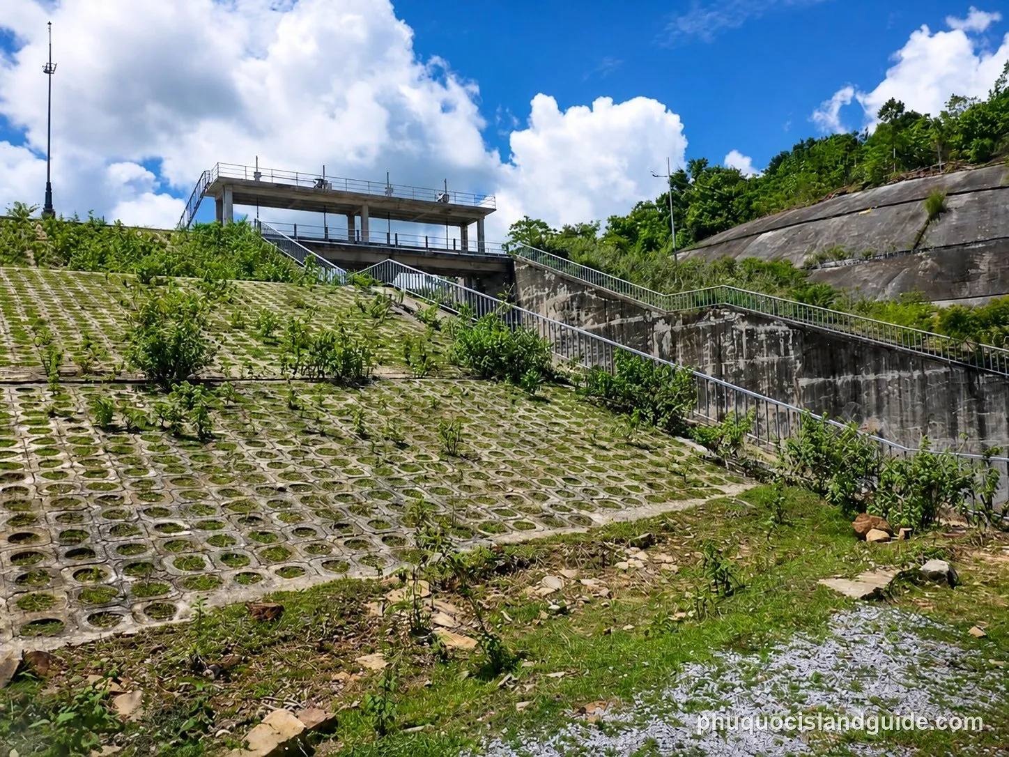 A concrete slope with grass growing through grid patterns, leading up to a concrete structure with stairs and railings. A green hill and blue sky are in the background.