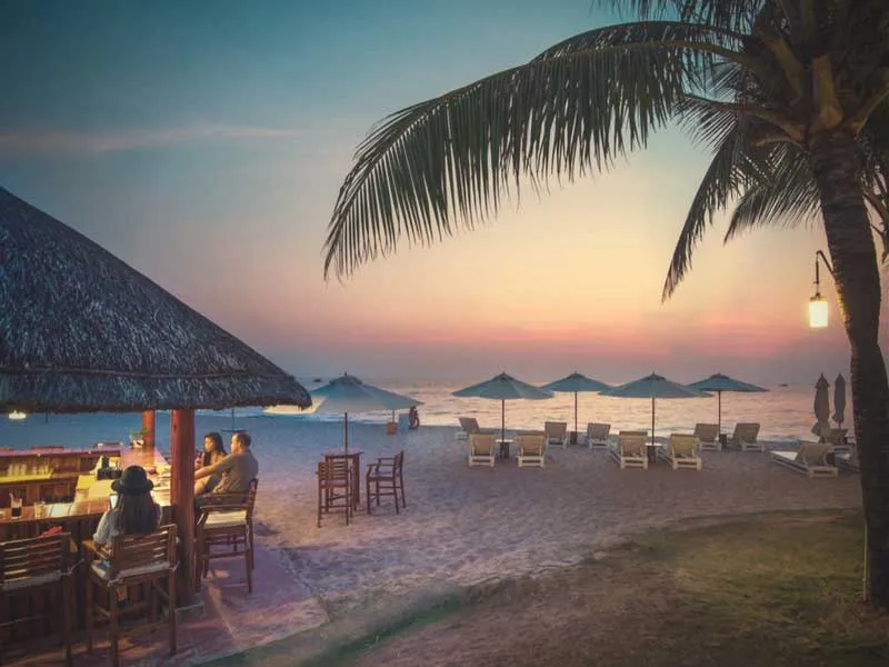 A tropical beach scene at sunset with a thatched-roof bar, people, palm trees, beach umbrellas, and lounge chairs on the sand by the ocean.