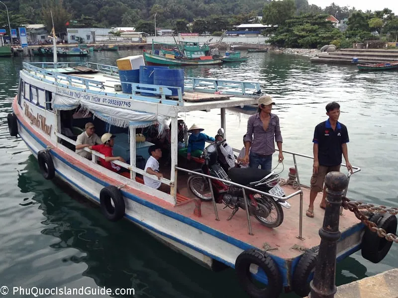A ferry boat with passengers and a motorbike docked at a pier in a busy harbor, with other boats and buildings in the background.