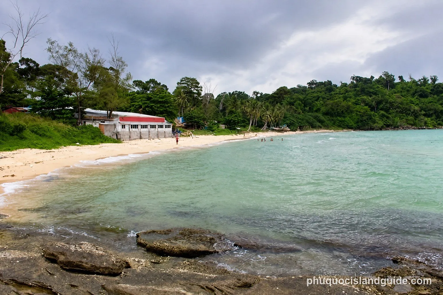 A tropical beach with clear turquoise water, a sandy shore, and lush green trees. A building with a red roof is visible on the left, and a few people are swimming.