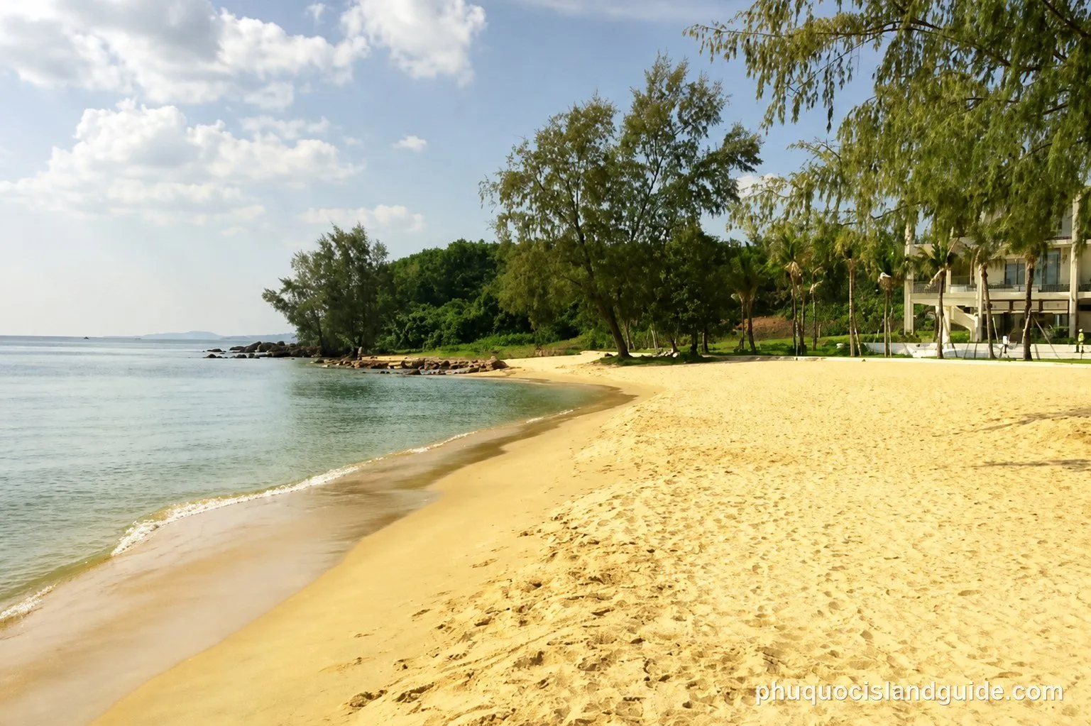 A sandy beach curves along a calm sea, bordered by lush green trees and some buildings in the distance under a partly cloudy sky.