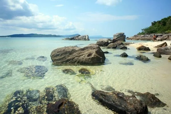 A clear, shallow tropical beach with large rocks in the water and on the sandy shore, with a distant island and blue sky with clouds.