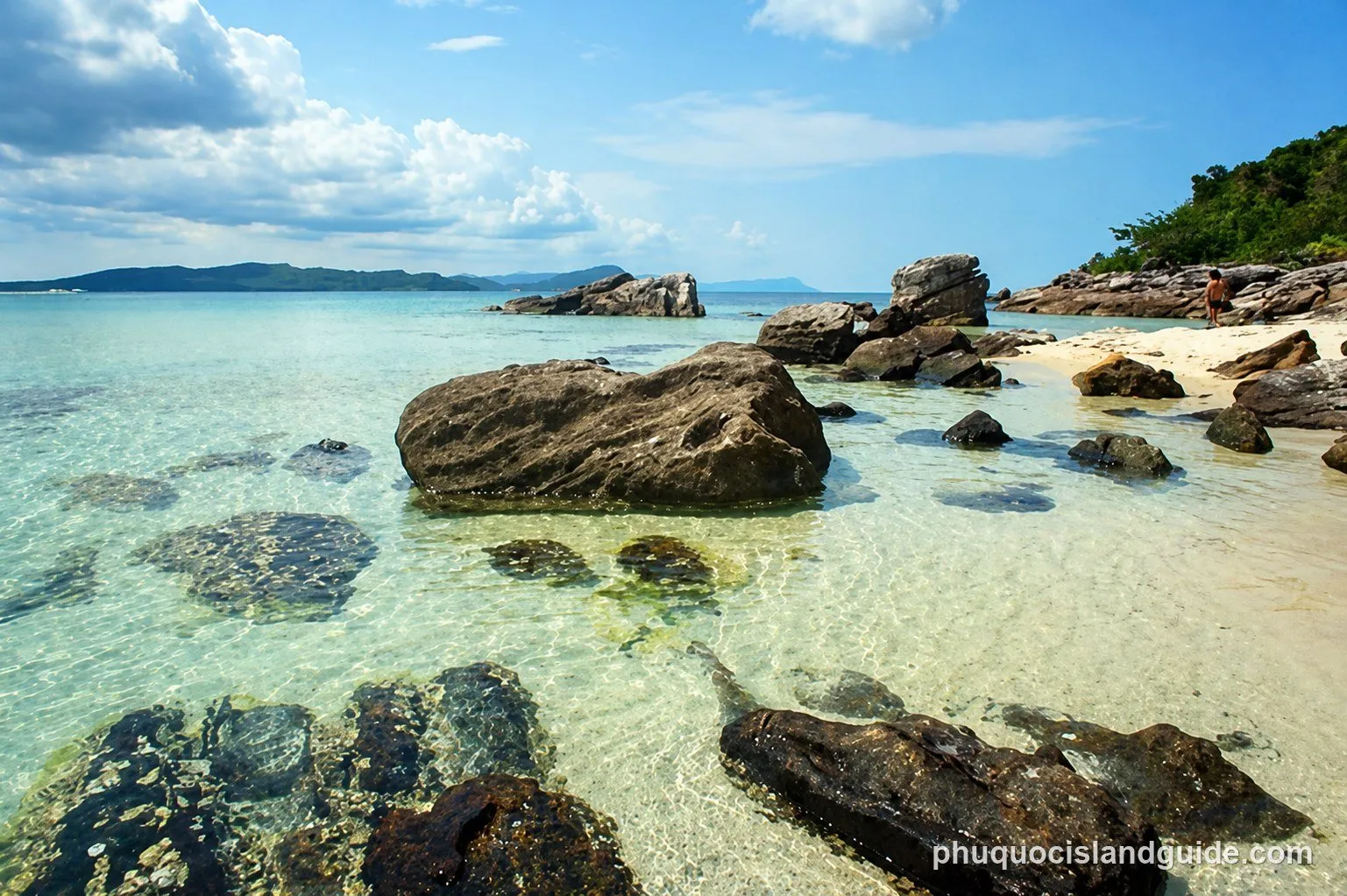 A clear, shallow tropical beach with large rocks in the water and on the sandy shore, with a distant island and blue sky with clouds.