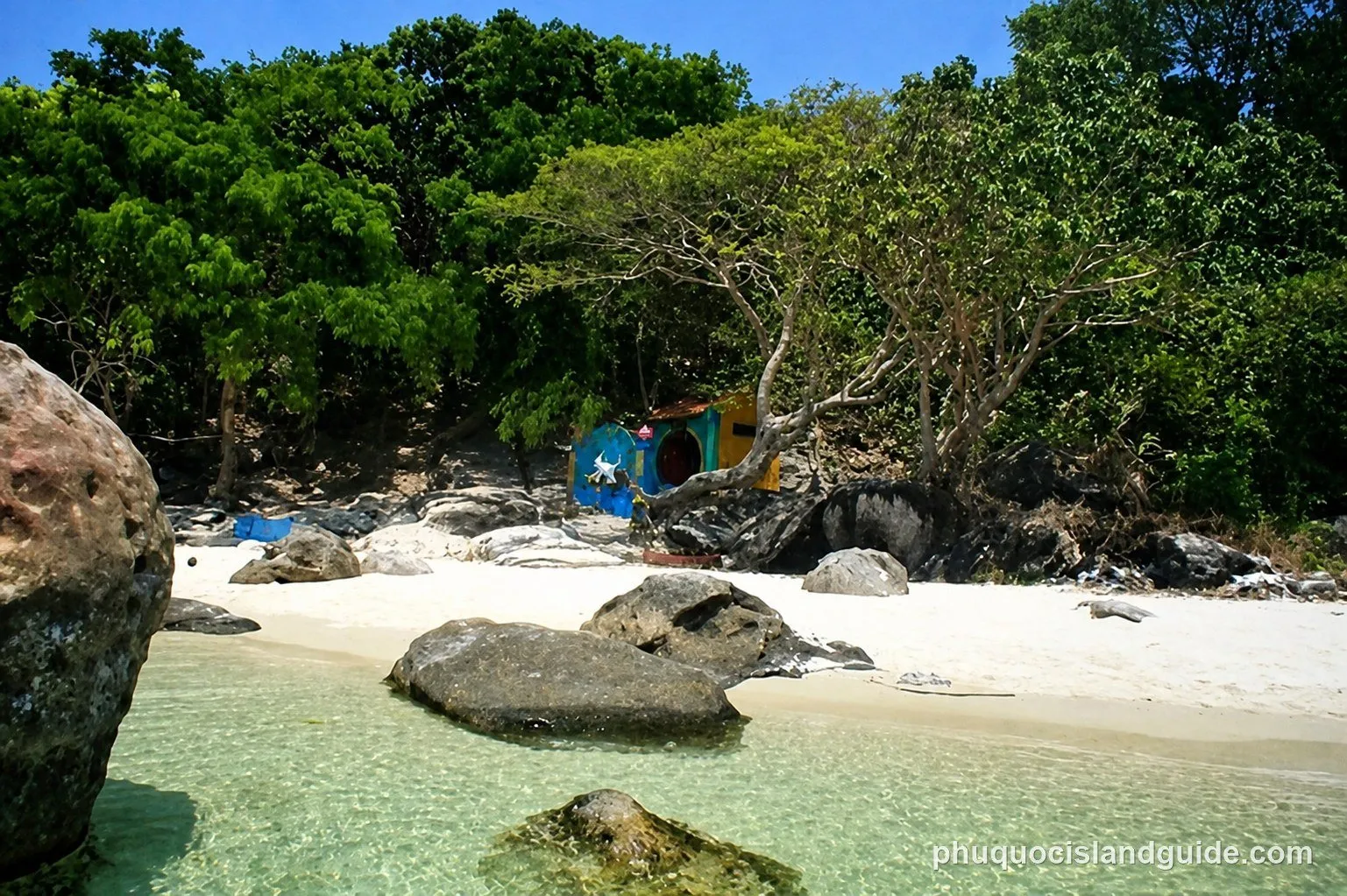 A tropical beach scene with clear turquoise water, white sand, and large rocks. Lush green trees line the shore, with a small, colorful hut nestled among them.