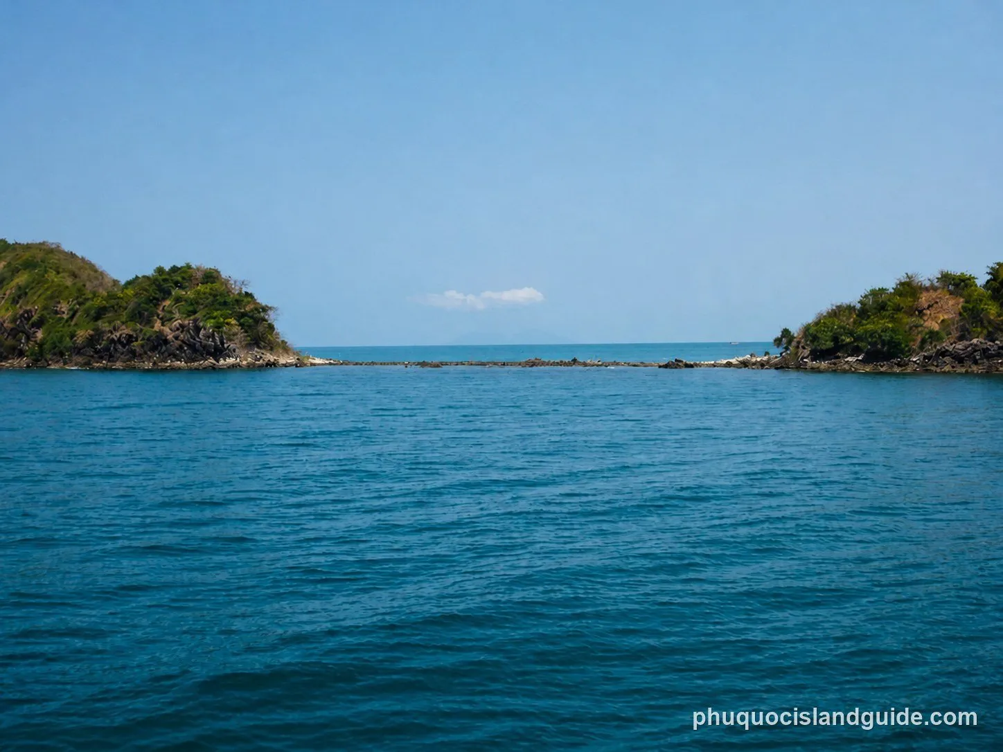Two small, rocky islands covered in green vegetation, separated by a narrow channel of blue sea and connected by a low-lying rocky bar under a clear blue sky.